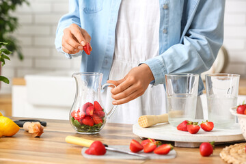 Woman putting strawberry into jug for lemonade in kitchen