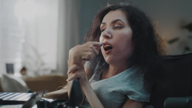 At home woman with disability in a wheelchair doing makeup at the table with cosmetics, in front of a mirror and applying a lip liner