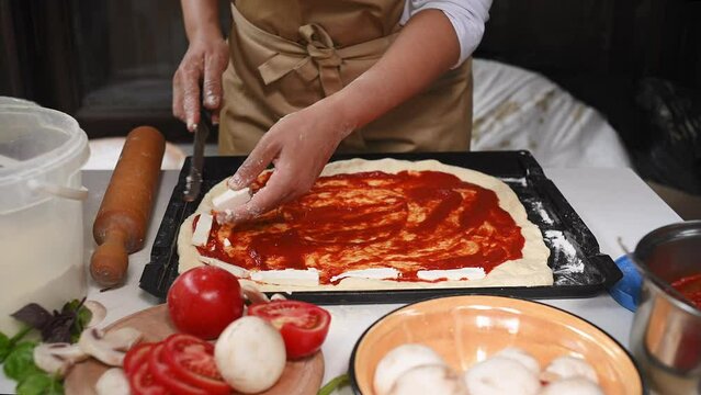 Close-up chef pizzaiolo makes homemade Italian pizza with tomato sauce, fresh mushrooms, feta cheese and basil leaves in a rustic kitchen. Traditional Italian cuisine. Healthy eating. Food consumerism
