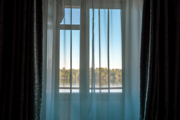 Bedroom window with curtains on the background of the lake and the forest, illuminated by the morning sun.