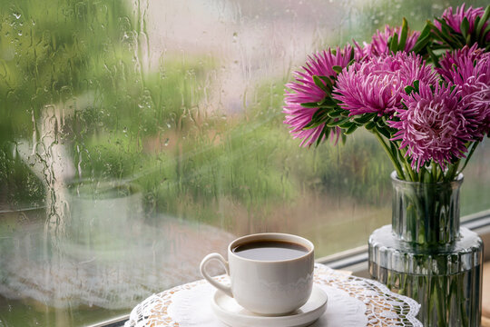 A cup of coffee and flowers in the vase standing next to a rainy window. 