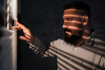 Young hipster male with beard looking through blinds he is spying on someone. Suspicious male...
