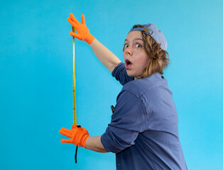 An adult woman in a shirt and a cap measures with a construction tape measure on an isolated blue background. Portrait of a cheerful woman in orange gloves. woman builder
