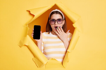 Indoor shot of shocked surprised woman wearing striped t shirt looking through breakthrough of...