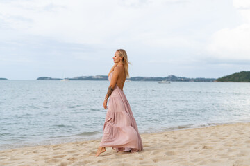 Beautiful young blonde woman in a long pink dress on the beach, tender, posing, the wind develops the dress hair, romantic, free