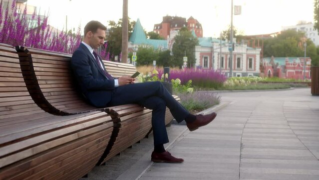 Young businessman in official dark blue suit sits on bench in park before meeting with partners and preparing to make presentation. Office clerk at lunch break working on cell or smartphone outdoors.