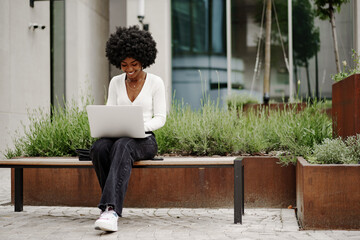 Young african american businesswoman working using laptop sitting on the bench in the city - Powered by Adobe