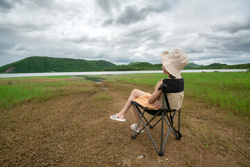 The young woman sits on the camping chair and looks at the view of the reservoir at the Huai Phak Reservoir, Tha Yang District, Phetchaburi, Thailand.