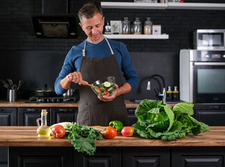 Smiling  man cooking salad, cutting fresh vegetables in modern black kitchen, happy satisfied male preparing vegetarian  food for dinner. Cabbage, tomatoes, zucchini, pepper on the back.