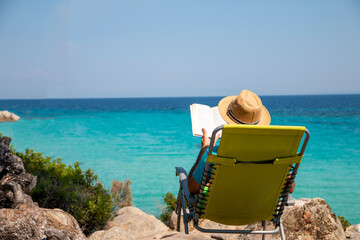 Fototapeta premium Portrait of a handsome man lying on a deckchair and reading a book. Happy mature man next to the beach on Aegean sea with beautiful view. Summer vacation concept.