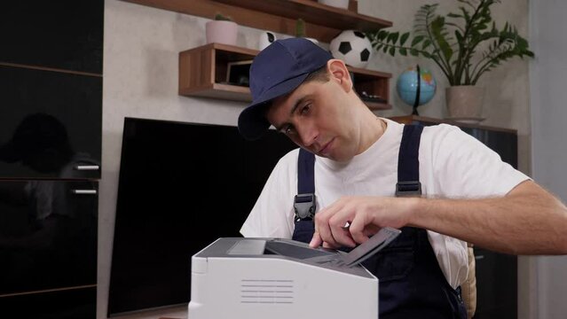 A male handyman in blue overalls inspects a printer before starting repairs.