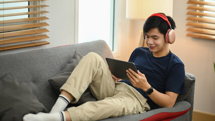 Satisfied young asian man using digital tablet, chatting in social networks while sitting on couch in cozy living room