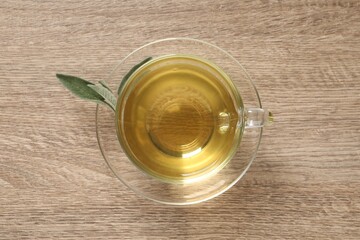 Cup of aromatic sage tea and fresh leaves on wooden table, top view