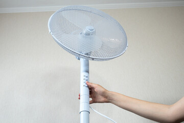 Woman is turning on Metal white modern fan or vent. Working electric floor standing fan in the...
