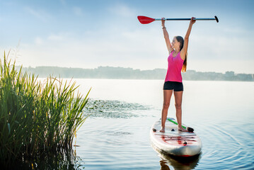 A beautiful woman holds an oar above her head while standing on a paddle board - obrazy, fototapety, plakaty