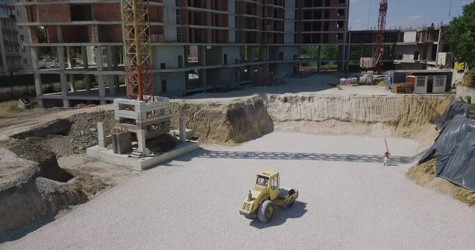 image from above on a construction site where a yellow tractor levels and compacts the new asphalt against the background of high-rise buildings under repair