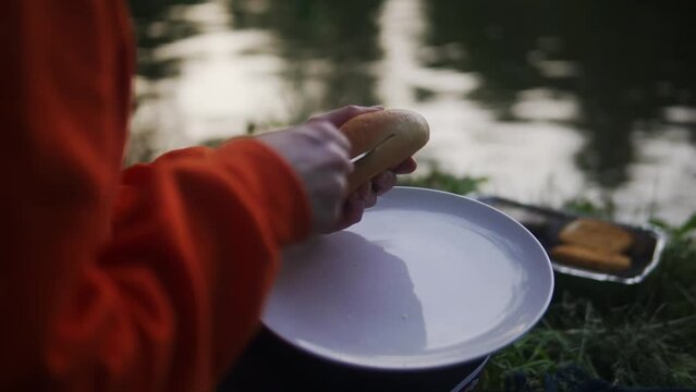A close up shot of a lady wearing full sleeve dress cutting burger bun while sitting along side of a pond