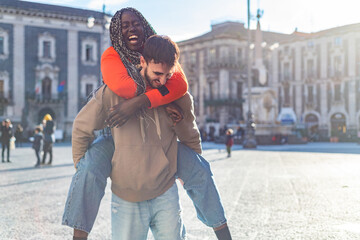 Smiling diverse young boyfriend carrying girlfriend on his back and laughing outdoors - boyfriend giving piggyback ride to girlfriends - Young multiracial couple having piggyback – happy multi ethnic  - Powered by Adobe