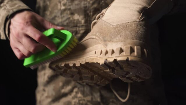 Soldier in camouflage uniform cleans his army shoes  with a green shoe brush in the dark room, close up. Taking care of military shoes at war.  Evening ritual of a military serviceman.