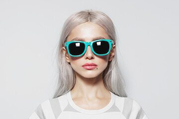 Studio portrait of serious, beautiful young girl with blonde hair in striped shirt on white background, wearing blue sunglasses.