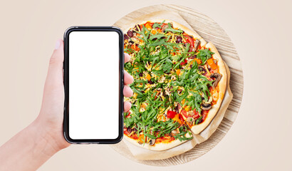 Close-up of male hand holding smartphone with empty blank on screen, on background of pizza isolated on pastel beige background. Vegan food. Delivery concept.