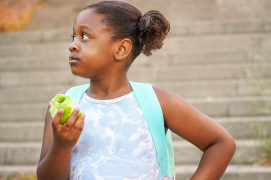 Elementary girl with backpack eating apple at the school entrance. healthy eating for children concept