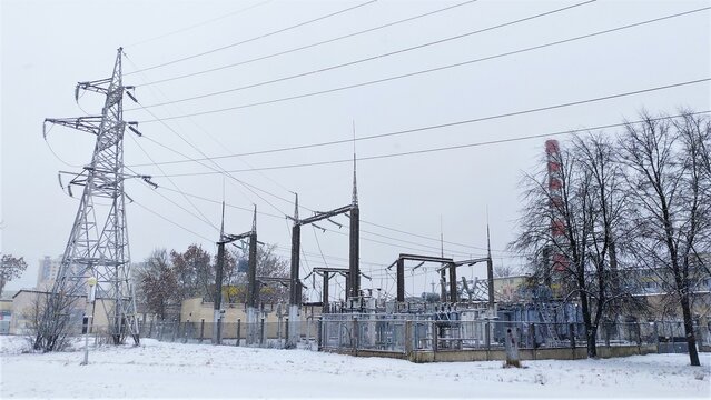 A transformer substation with a metal power line tower, ceramic insulators, and lightning rods is located on a snow-covered lawn and surrounded by a fence. Nearby are a factory chimney and buildings. 