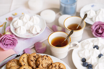 two cups of tea on table with chocolate cookies, meringue, candles, pink rose and book