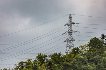 Pylon with electric cables between trees in a rural area against cloudy sky