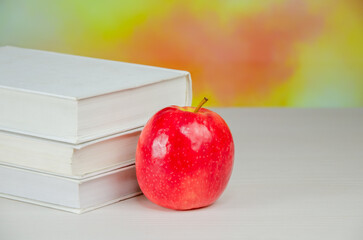 Juicy fresh apple stands on desktop next to stack of books over blurry autumn background.