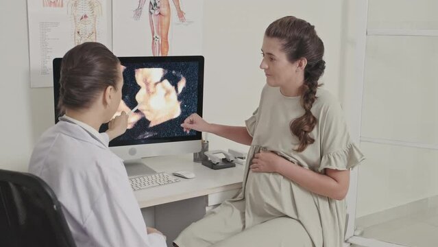 Female doctor showing ultrasound image of baby in womb on computer screen and talking to pregnant woman during prenatal consultation in hospital