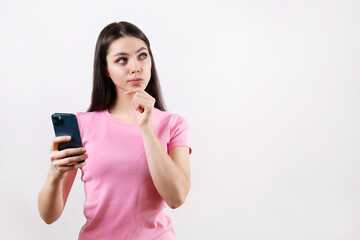 Fototapeta premium Young beautiful woman with thoughtful facial expression holding a blank screen phone. Portrait of female model with cellphone calculating in her mind. Close up, copy space, isolated white background.