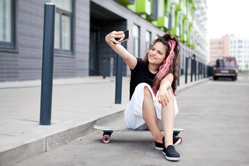Fototapeta premium Teen girl sitting on road on skateboard, taking selfie on her phone and smiling. Lifestyle youth culture. Trendy teen with long pink colored hair
