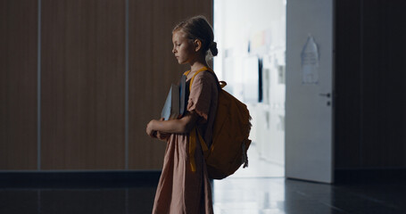 Worried little schoolgirl stop in school hallway alone. Student holding books. - obrazy, fototapety, plakaty