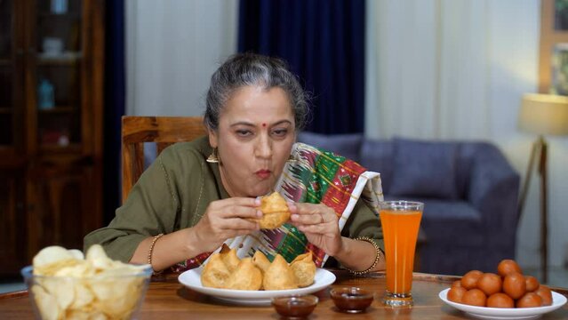 An aged Indian housewife eating Samosa - Indian street food  deep-fried  stuffed samosa  unhealthy food  trans fat  oily food. An old Indian lady bingeing on unhealthy food - poor lifestyle  cheat day