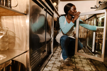 Young african woman squatting and putting bottles out the fridge