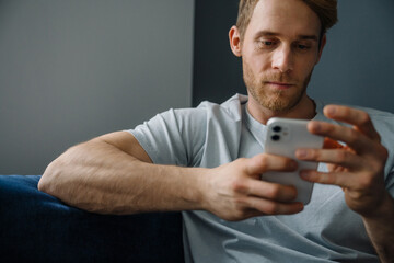 Young bearded man using cellphone while sitting on sofa at home