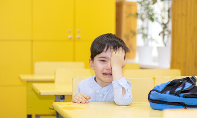 preschooler boy is crying sitting on school desk. angry unhappy sad upset kid don't want to go back...