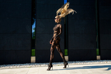 High fashion look. Glamour stylish model in black latex clothing and and high heels on is walking in the city. Sexy  blonde woman in bodystocking lingerie is posing outdoor. Sexy Lingerie model. - obrazy, fototapety, plakaty