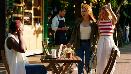 Two beautiful women are walking in a park towards their friend who is wearing red wireless headphones and sitting at the table