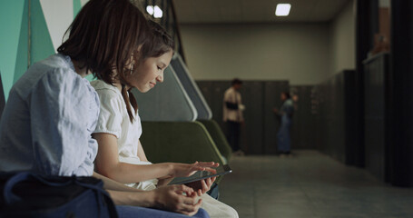 Smiling schoolgirls using tablet at school break. Two teens talking at lockers.