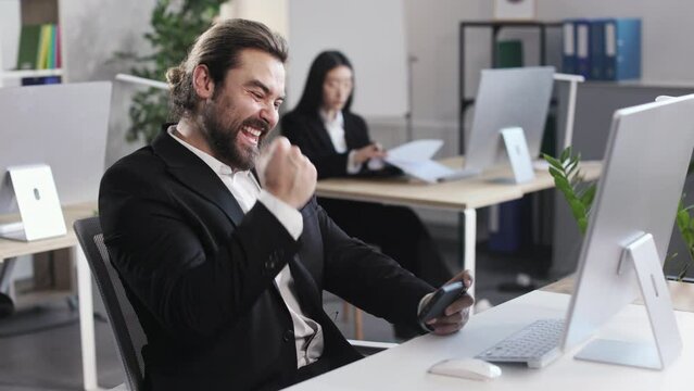 Cheerful young man in stylish black suit playing games on smartphone at workplace. Relaxed office manager making yes gesture during victory. Entertainment and relax at office.