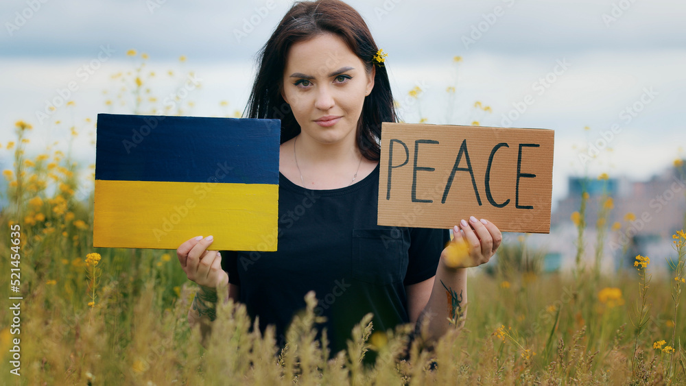 Wall mural Unhappy lady brunet woman activist pick up two paper banners with inscription peace cardboard in blue yellow ukrainian flag colors sad young girl stand outside protesting military conflict in Ukraine