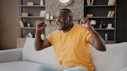 Senior football fan african american man bachelor sit on cozy sofa in apartment watch TV sport...