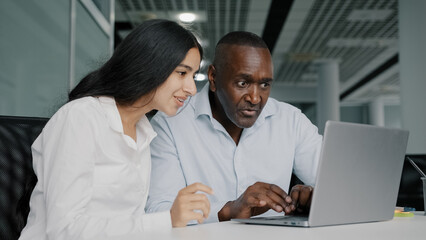 Two diverse colleagues businesspeople african senior man lawyer and arabian woman worker sit in modern co-working office look at laptop consult financial insurance negotiating discuss startup project