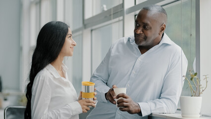 Two diverse age multi-ethnic colleagues businesspeople co-workers partners friends african senior adult man arab young woman standing indoors talk drink coffee tea have pleasant informal conversation
