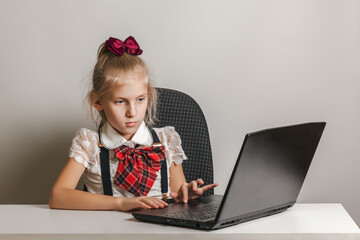 A little girl in a school uniform uses a laptop at a table on a white background