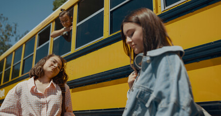 Two cheerful girls talking at school bus. Schoolboy looking out bus window.