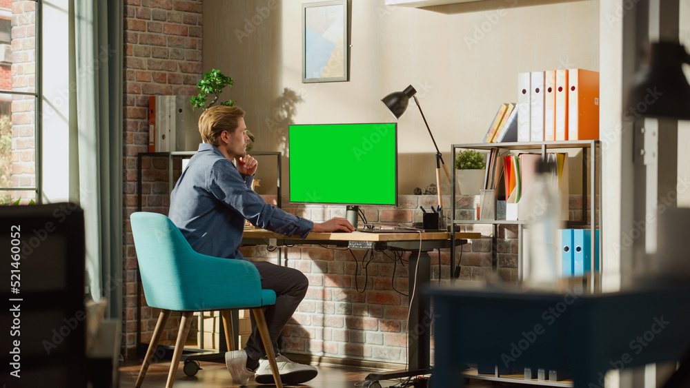 Poster Young Handsome Man Working from Home on Desktop Computer with Green Screen Mock Up Display. Male Checking Corporate Accounts, Messaging Colleagues. Loft Living Room with Big Window.