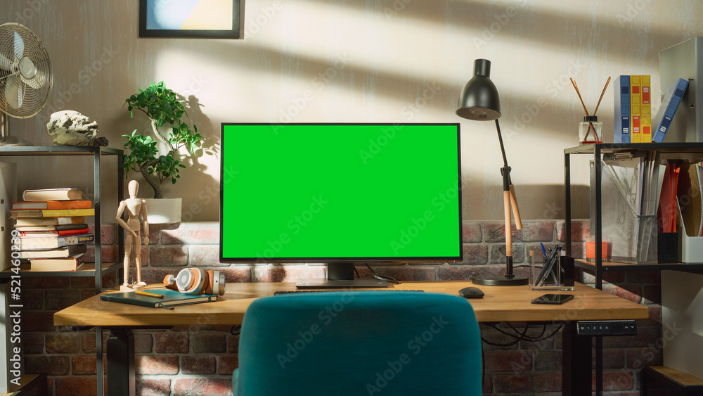Poster Desktop Computer Monitor Standing on a Table with a Green Screen Chromakey Mock Up Display. Cozy Empty Loft Apartment with a Lamp, Notebooks, Smartphone and Headphones on the Table.